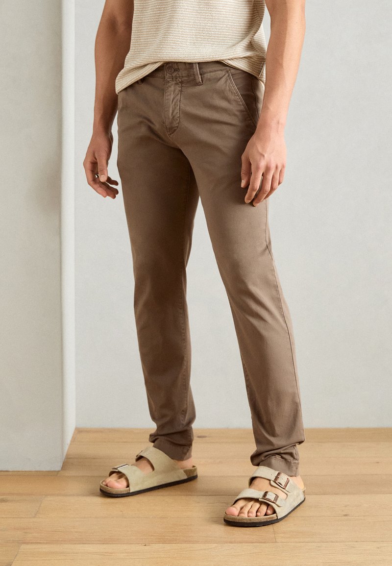 Pantaloni slim-fit marroni in cotone, con vita con bottone e tasche frontali, abbinati a sandali aperti di colore chiaro.