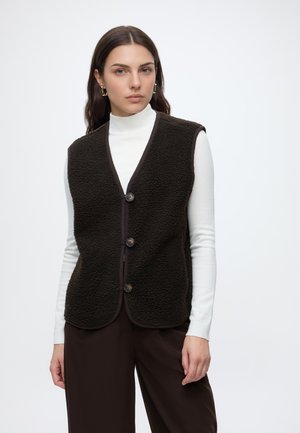 Femme aux longs cheveux foncés portant un col roulé blanc, un gilet polaire noir boutonné, un pantalon brun foncé et des boucles d’oreilles géométriques dorées, debout sur fond blanc.