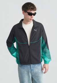 Jeune homme portant une veste zippée Puma noire et turquoise, un t-shirt blanc, un jean bleu clair imprimé Puma et des lunettes de soleil noires, regardant sur le côté.