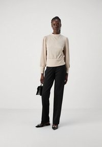 Beige, laskostettu pitkähihaisen yläosa, jossa on ribbineuloshelma, yhdistetty mustiin suoralahkeisiin housuihin. Mustat tasapohjaiset kengät ja pieni teksturoitu käsilaukku.