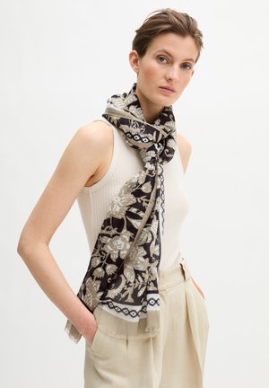 Vrouw met een mouwloze witte top en beige broek, met een zwarte en beige bloemen sjaal om haar nek geknoopt, handen in de zakken, neutrale achtergrond.