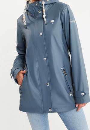 Blå vandtæt hættetrøje regnjakke med knaplukning, snøre i hætten og sidelommer, båret med lyseblå jeans.