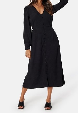 Woman wearing a long-sleeve black midi dress with front buttons and black open-toe heeled sandals on a white background.