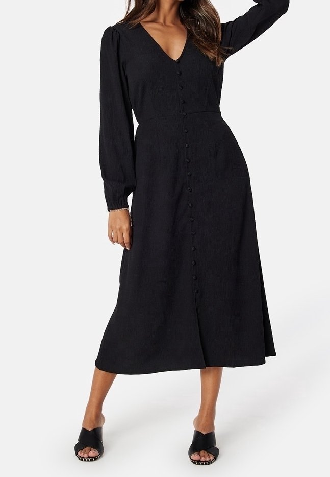 Femme portant une robe midi noire à manches longues avec des boutons à l'avant et des sandales à talons ouvertes noires sur un fond blanc.