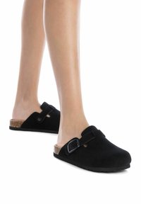 Zuecos de ante negro con punta redondeada, suela de corcho y una correa negra ajustable. Presentan una textura suave y un diseño minimalista.