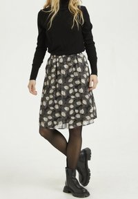 Jupe en tissu noir à motifs de feuilles blanches, taille élastique, longueur genou, superposée à un jupon noir uni, associée à des bottines noires.