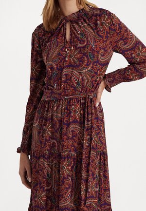 Mujer que lleva un vestido morado de manga larga con un patrón paisley rojo y verde, escote con abertura en forma de ojo, y cinturón de tela a juego atado a la cintura.