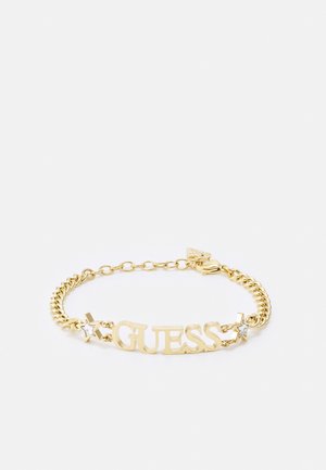 Guld kæde armbånd med "GUESS" bogstaver og stjerne vedhæng på hver side, hvoraf det ene har en lille klar sten, på en hvid baggrund.