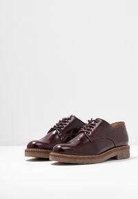 Bordeauxfarbene Lederschuhe mit glänzender Oberfläche, runder Zehenform, Schnürdesign und einer hellbraunen strukturierten Sohle. Glattes Obermaterial mit minimaler Nahtverarbeitung.