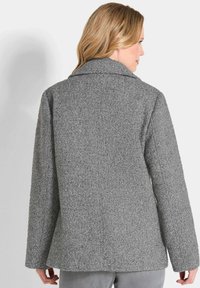 Femme aux cheveux blonds portant une veste en herringbone gris texturé, de dos contre un fond clair.