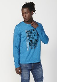 Blue long-sleeve T-shirt made of textured fabric featuring a geometric skull graphic in black, with "ENGINE CUSTOM MOTOR RACING" text.