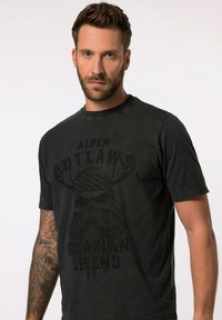 Sort kortærmet t-shirt med rund hals. Har et print af en skægget figur med horn og teksten "ALPEN OUTLAWS BAVARIAN LEGEND" i mørk design.