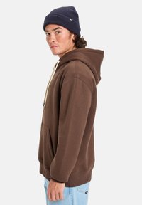 Sudadera con capucha marrón, con un bolsillo tipo canguro, cordones ajustables y puños acanalados, mostrada llevada con un gorro azul.
