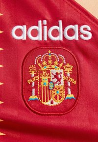 Κόκκινη αθλητική φανέλα με λευκό λογότυπο Adidas πάνω από λεπτομερή κεντημένο ισπανικό εθνόσημο που εμφανίζει ένα στέμμα και ασπίδα.