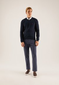 Marineblauwe gebreide trui met een V-hals, gecombineerd met lichtgrijze broek en bruine instapschoenen. Heeft een klein rood logo op de trui.