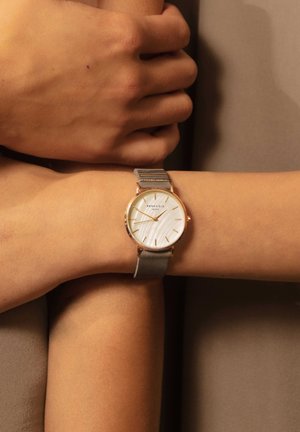 Montre argentée et dorée avec un cadran blanc texturé, dotée d'index en or et d'un bracelet en cuir gris.