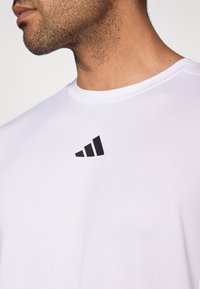 Hvid atletisk T-shirt med korte ærmer og rund halsudskæring i tekstureret stof. Har en sort trekantet logo på den øverste venstre del af brystet.