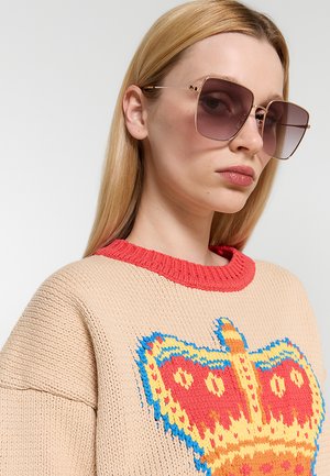 Donna bionda che indossa occhiali da sole quadrati oversize e un maglione beige con colletto rosso e motivo corona colorato.