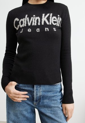 Kvinde iført en sort Calvin Klein Jeans sweater og blå cowboybukser, med den ene hånd hvilende i lommen.