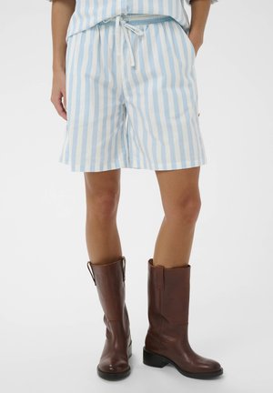 Person iført lyseblå og hvide shorts med lodrette striber, snøre i taljen og mørkebrune læderstøvler til midt på læggen på hvid baggrund.
