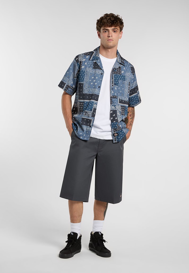 Camicia a maniche corte con fantasia in tonalità blu, modello a vestibilità comoda, abbinata a pantaloncini grigio scuro fino al ginocchio. Il modello indossa scarpe da ginnastica nere e calzini bianchi.