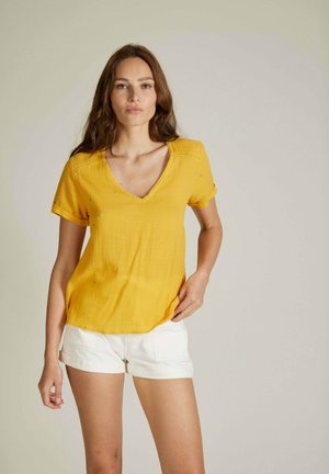 Haut jaune à manches courtes avec un col en V, présentant un tissu texturé et de petits éléments décoratifs. Associé à un short blanc retroussé.