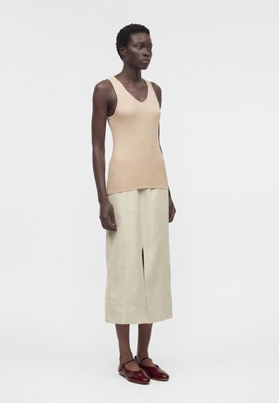 Γυναίκα στέκεται φορώντας μπεζ ριμπ τοπ χωρίς μανίκια, κρεμ midi φούστα με σκίσιμο μπροστά και μπορντό παπούτσια τύπου Mary Jane σε απλό φόντο.