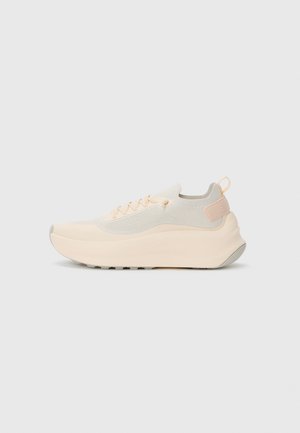 Laag profiel, lichtbeige sportshoe met gebreide bovenkant, dikke gevoerde zool, vetersluiting en trekband aan de hiel, weergegeven in zijaanzicht.