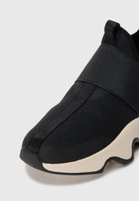Svart slip-on sneaker med pustende mesh-overdel, bred elastisk stropp, trekkhank på hælen og tykk beige såle med bølgete svart mønster.
