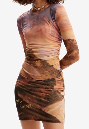 Frau trägt ein figurbetontes, langärmliges Kleid mit Rüschen in einem braunen und pinkfarbenen abstrakten Landschaftsprint.