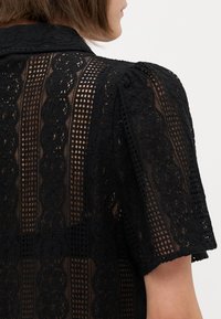 Top in pizzo nero con colletto, motivi floreali texture e maniche corte a sbuffo con una trama trasparente e aperta.