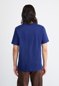 Persoană cu păr întunecat de lungime medie, purtând un tricou simplu albastru cu mânecă scurtă și pantaloni maro, întorcându-se cu fața la un fundal alb.