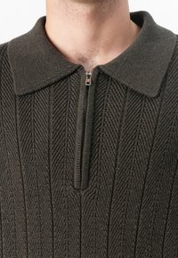 Κοντινό πλάνο ενός ατόμου που φοράει ένα σκούρο πράσινο πλεκτό πουλόβερ με γιακά και έναν ανοιχτόχρωμο μεταλλικό φερμουάρ που είναι μερικώς κλειστό μπροστά.