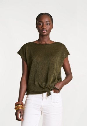 Femme portant un haut vert olive à manches courtes noué à la taille, un pantalon blanc et plusieurs bracelets en or et en bois à son poignet gauche.