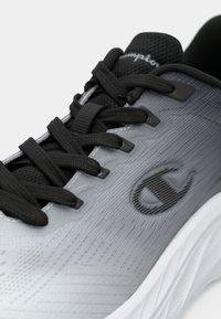 Γκρι αθλητικό παπούτσι με υφασμάτινη επιφάνεια, μαύρα κορδόνια, λογότυπο στο πλάι και λευκή σόλα. Ο σχεδιασμός διαθέτει φινίρισμα με διαβάθμιση χρώματος.