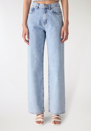 Jeans azzurri chiari a vita alta con gambe larghe su una persona in piedi con sandali bianchi con cinturini, corti in vita, sfondo neutro.