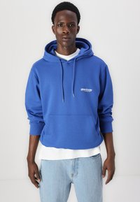 Sudadera azul con capucha y bolsillo tipo canguro, con cordones y detalle de logotipo blanco, superpuesta sobre una camiseta blanca, combinada con jeans de mezclilla claros.