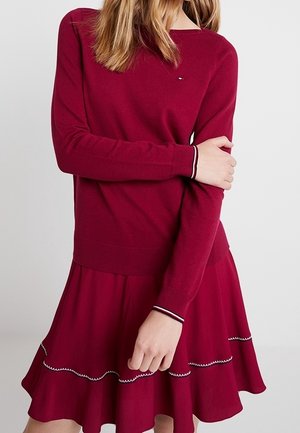 Kvinde iført en bordeaux langærmet sweater med et matchende skørt med hvidt zigzagbånd langs den flæsebelagte kant.