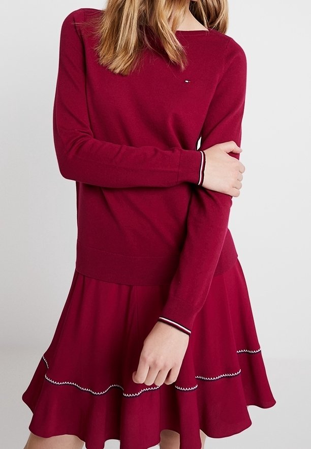 Femme portant un pull bordeaux à manches longues avec une jupe assortie ornée d'un galon zigzag blanc le long de l'ourlet volanté.