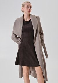 Beiger Wollmantel mit breitem Kragen, kombiniert mit einem dunkelbraunen Cordkleid. Der Mantel hat einen einzelnen Knopf und lange Ärmel.