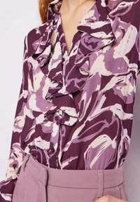 Blusa viola con motivo floreale astratto in crema e viola chiaro, con ruffles lungo il collo e chiusura con bottoni. Tessuto morbido.