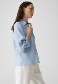 Camicia azzurro chiaro con texture, con bottoni frontali e maniche arrotolate, caratterizzata da una vestibilità rilassata e fessure laterali, abbinata a pantaloni bianchi.