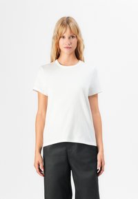 Donna bionda che indossa una semplice t-shirt bianca a maniche corte e pantaloni neri, in piedi contro uno sfondo bianco, rivolta in avanti.