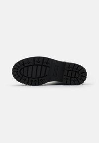 Svart gummisul med djupt mönster, med ribbor och cirkulära detaljer för grepp. Undersidan är slät med varumärkesdetalj.