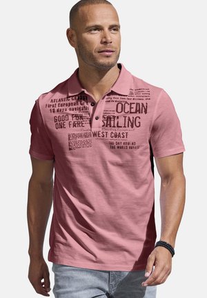 Mann trägt ein rosa Poloshirt mit schwarzem, segelbezogenem Text und Grafiken auf der Brust, kombiniert mit hellgrauen Jeans und einem schwarzen Perlenarmband.