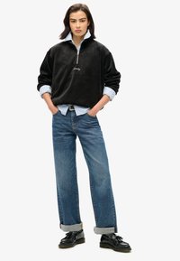 Pullover in pile nero con zip e dettagli ricamati, indossato sopra una camicia azzurro chiaro. Abbinato a jeans blu a vita alta e a gamba larga.