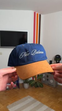 Boné azul com pala castanha, com texto bordado branco "New Balance" na frente. Material de algodão com ilhós de ventilação metálicos.