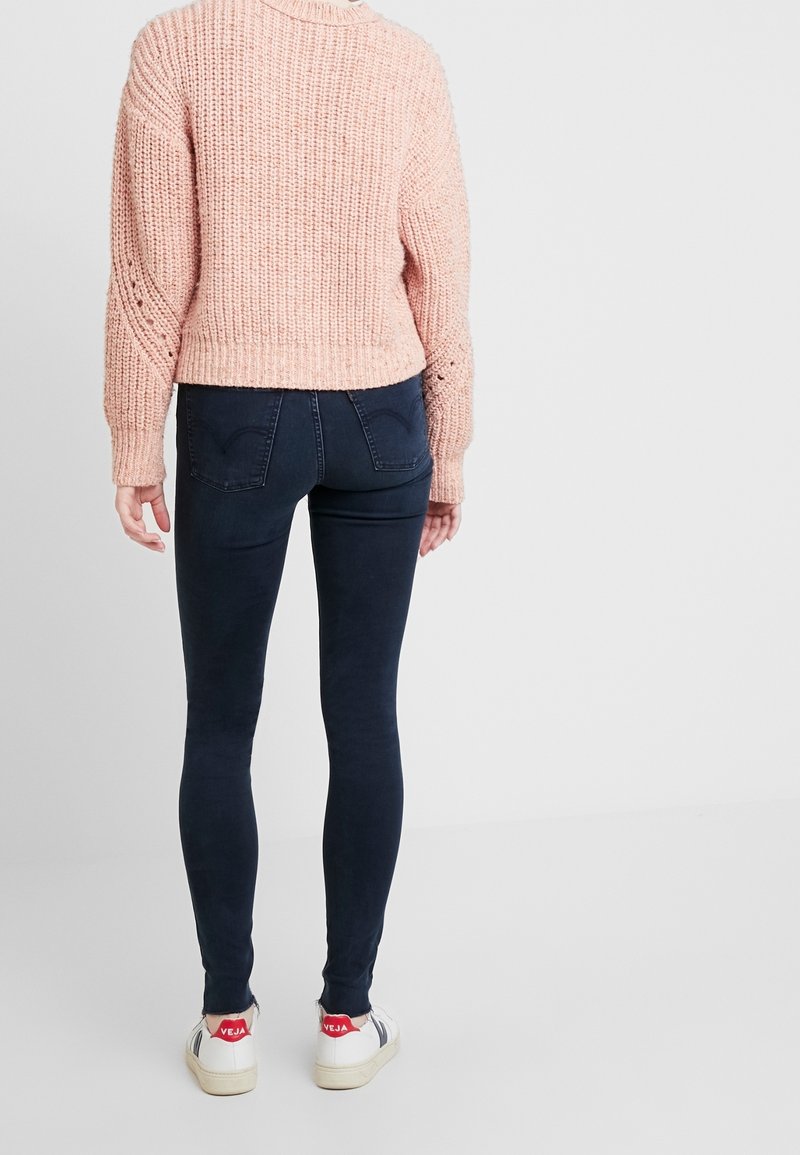 Suéter grueso rosa con corte corto, combinado con jeans ajustados azul oscuro y zapatillas blancas con acentos rojos en el talón.