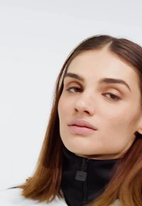 Zwarte rits-jack met kraag en een gladde textuur, gecombineerd met lang, steil bruin haar; subtiele make-up; neutrale achtergrond.