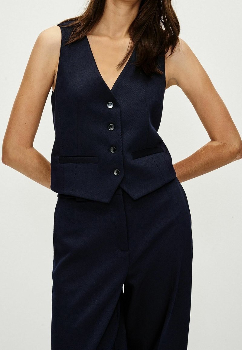 Femme portant un gilet bleu marine sans manches à quatre boutons et un pantalon taille haute assorti, debout les mains derrière le dos.